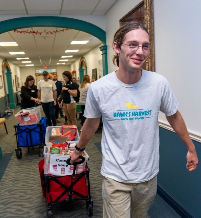 A Hawk’s Harvest volunteer pulls a wagon filled with donated food through a UNCW hallway as other volunteers work in the background.