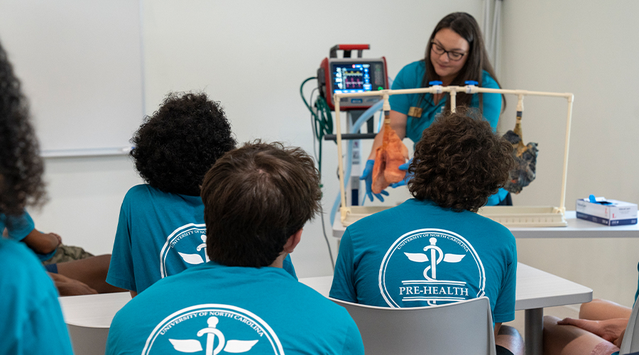 Students observe an instructor demonstrating medical procedures on a training mannequin in a clinical simulation room.